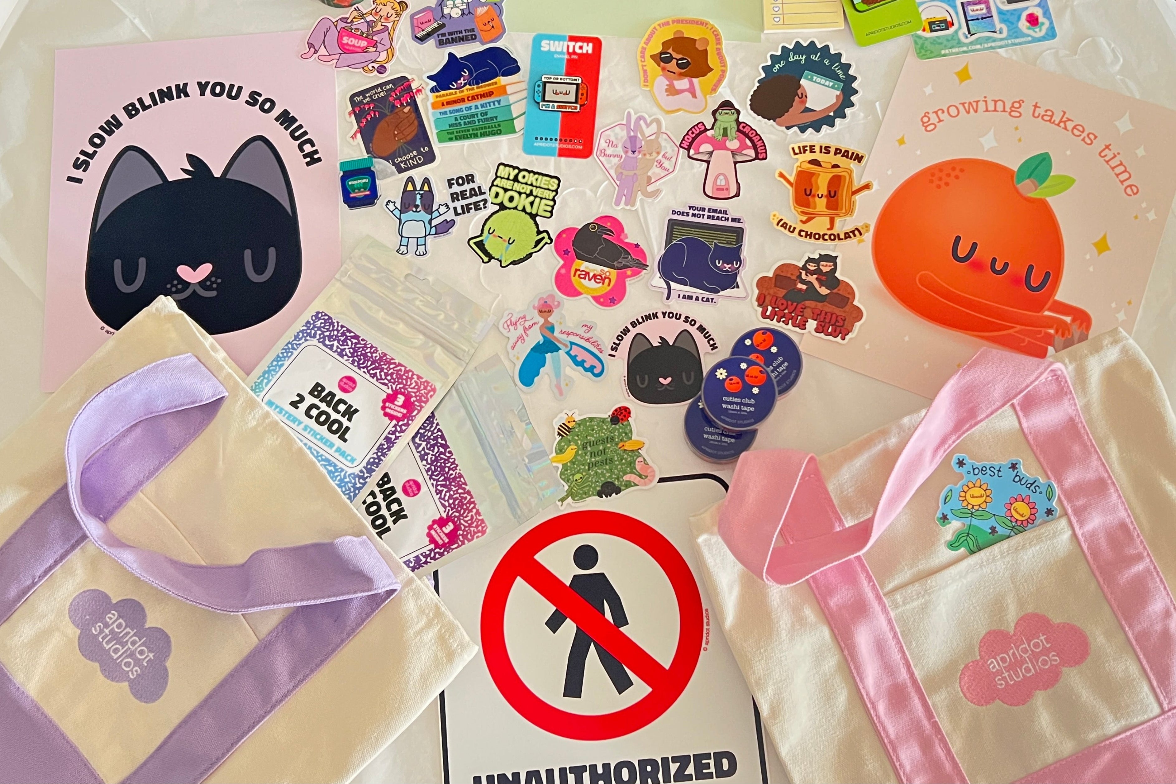A collection of cute pins, stickers, and washi tape, prints, and mystery sticker bags arranged on a white table runner. The bags feature pastel pink, lavender, and cream colors with a cloud 'apridot studios' logo. 
 
